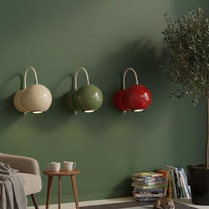May include: Three modern wall sconces in cream, olive green, and red, each with a spherical shade and a curved, white metal arm. The sconces are mounted on a dark green wall. A small side table, chair, and a potted tree are in the foreground.