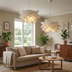 May include: A living room scene featuring a beige sofa, wooden furniture, and two white, cloud-like pendant lights. Green plants in terracotta pots add a natural touch. A round wooden coffee table sits in front of the sofa.