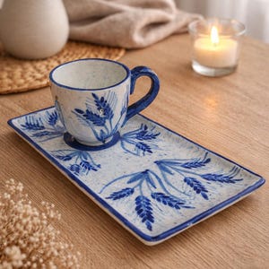 Könnte beinhalten: Ein Keramik-Teebecher und ein rechteckiges Untertassen-Set. Die Tasse und die Untertasse sind weiß mit blauen Blumendesigns und einem blauen Rand. Die Tasse hat einen blauen Griff. Das Set steht auf einem Holztisch.