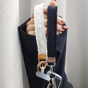 May include: Two wrist straps, one white and one black, with floral lace patterns. The straps have gold-colored metal hardware and are attached to a set of keys and a phone. The background is a neutral color.