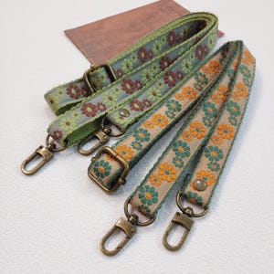 Puede incluir: Tres correas decorativas para bolsos con diseños florales. Una correa es verde con flores marrones y verdes, otra es beige con flores naranjas y verdes, y la tercera es verde con flores marrones y verdes. Cada correa tiene herrajes metálicos.