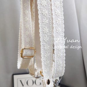 Puede incluir: Primer plano de una correa de bolso color crema con un diseño de encaje floral. La correa presenta herrajes metálicos dorados, que incluyen una hebilla rectangular y un anillo circular. El texto "ZiYuan original design" es visible.