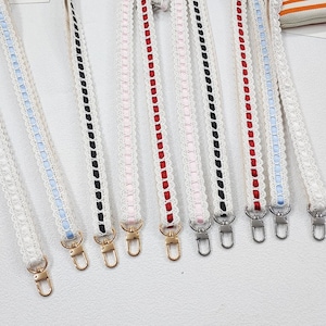 Op de afbeelding: Een verzameling witte kanten bandjes met gekleurde lintaccenten, waaronder rood, zwart, roze en blauw. Elke band heeft een metalen sluiting aan het uiteinde, geschikt om aan een tas te bevestigen. De bandjes worden tegen een witte achtergrond getoond.