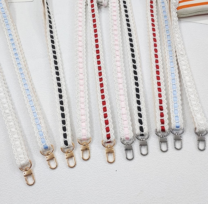 Op de afbeelding: Een verzameling witte kanten bandjes met gekleurde lintaccenten, waaronder rood, zwart, roze en blauw. Elke band heeft een metalen sluiting aan het uiteinde, geschikt om aan een tas te bevestigen. De bandjes worden tegen een witte achtergrond getoond.