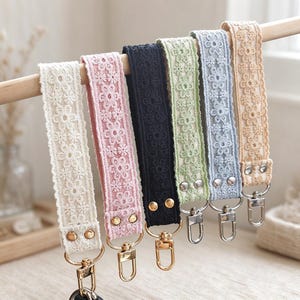 May include: Six fabric wristlet keychains featuring a floral lace overlay in cream, pink, black, sage green, light blue, and beige. Each strap is secured with two metal rivets and includes a swivel lobster clasp for attaching keys or accessories.