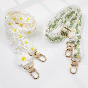 May include: Two decorative purse straps. One is white with yellow daisy accents and gold-colored clasps. The other is white with green daisy accents and gold-colored clasps. Both straps are made of lace.