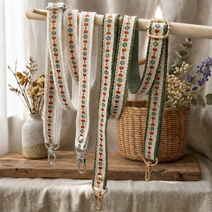 May include: Two adjustable bag straps featuring a cream-colored woven base with a repeating pattern of small orange hearts and green floral motifs. Each strap includes gold-toned metal swivel lobster clasps and rectangular sliders for length adjustment.