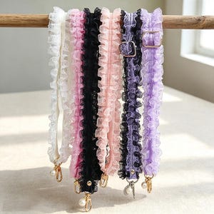 Könnte beinhalten: Eine Kollektion von sechs gerüschten Taschenriemen aus Spitze in Weiß, Pink, Schwarz, Hellrosa, Dunkellila und Helllila. Jeder Riemen hat einen gerüschten Stoffrand, eine goldfarbene verstellbare Schnalle und einen goldfarbenen Karabiner mit Perlenanhänger.