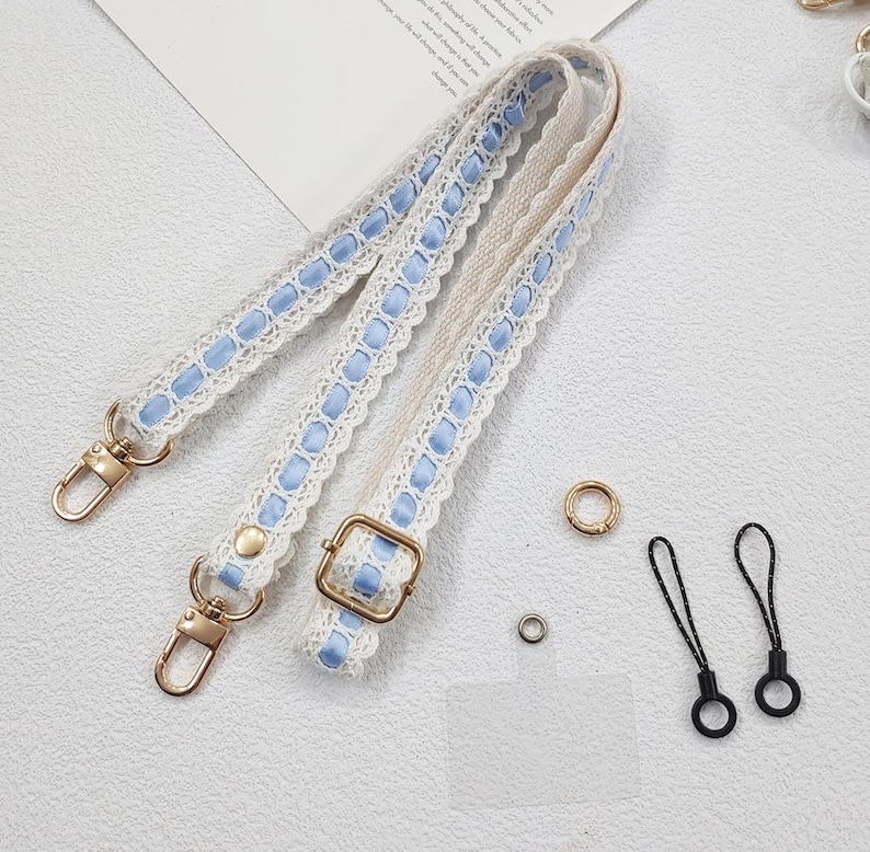 Op de afbeelding: Een cr&egrave;mekleurige tasriem met blauwe lintaccenten en goudkleurige hardware. De riem heeft een delicate kanten rand en een gesp voor verstelling. Ook inbegrepen zijn kleine gouden ringen en zwarte lussen.