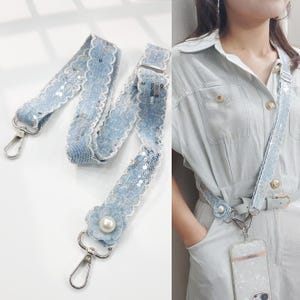 May include: A light blue denim phone strap with white lace trim and silver sequins. The strap has silver metal clasps and a decorative flower with a pearl center. The strap is shown on a person wearing a light blue outfit.
