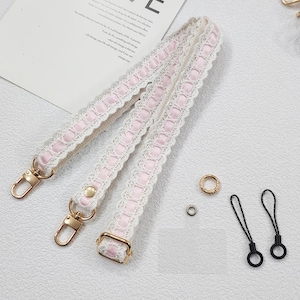 Op de afbeelding: Een cr&egrave;mekleurige handtasriem met roze lint en witte kanten rand. De riem heeft goudkleurige metalen sluitingen en hardware. Extra kleine gouden en zwarte accessoires zijn ook inbegrepen.