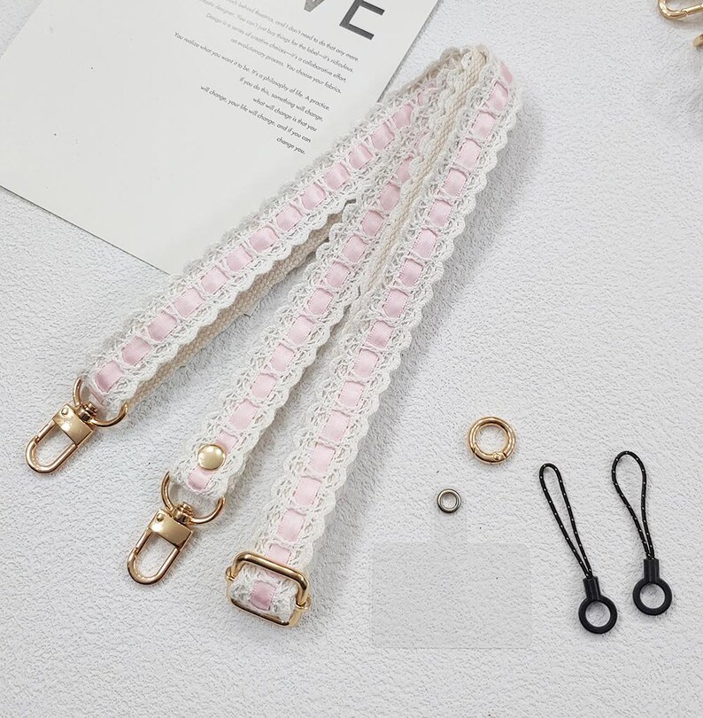 Op de afbeelding: Een cr&egrave;mekleurige handtasriem met roze lint en witte kanten rand. De riem heeft goudkleurige metalen sluitingen en hardware. Extra kleine gouden en zwarte accessoires zijn ook inbegrepen.