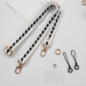 Op de afbeelding: Cr&egrave;mekleurige tasriem met zwarte lintaccenten en goudkleurige hardware. De riem heeft een delicaat kanten ontwerp. Extra gouden ringen en zwarte telefoonbedels zijn ook zichtbaar.