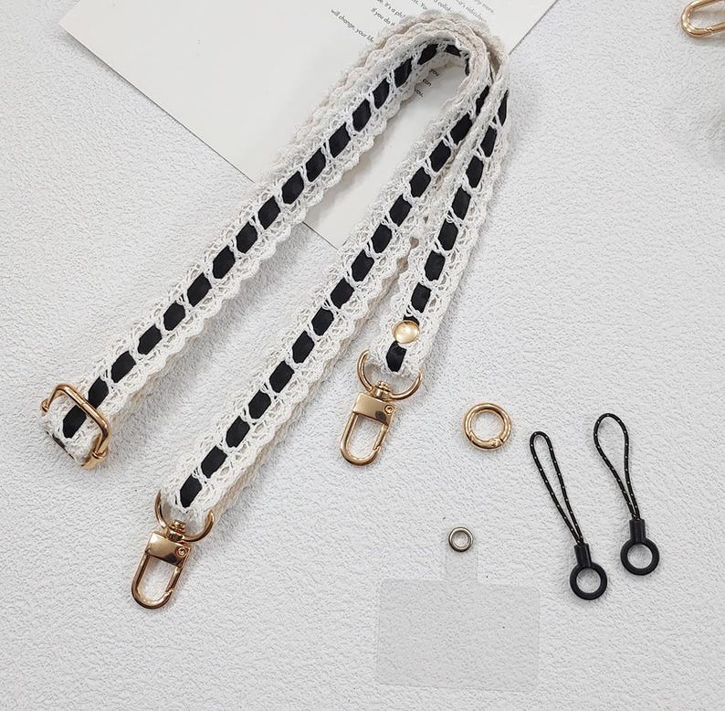 Op de afbeelding: Cr&egrave;mekleurige tasriem met zwarte lintaccenten en goudkleurige hardware. De riem heeft een delicaat kanten ontwerp. Extra gouden ringen en zwarte telefoonbedels zijn ook zichtbaar.