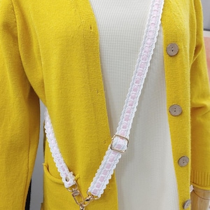 Op de afbeelding: Een gele cardigan met houten knopen en een wit en roze patroonband. De band is bevestigd aan een telefoonhoesje. De cardigan zit over een wit shirt.