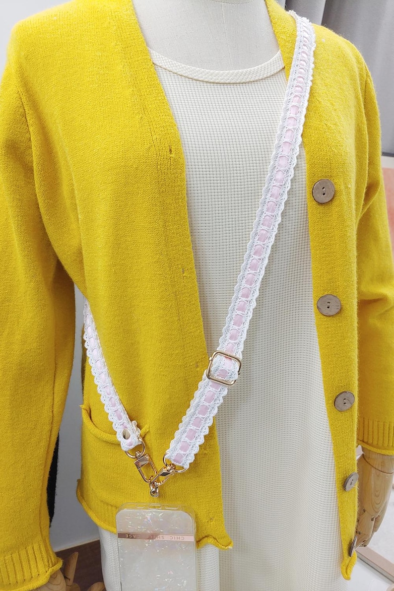 Op de afbeelding: Een gele cardigan met houten knopen en een wit en roze patroonband. De band is bevestigd aan een telefoonhoesje. De cardigan zit over een wit shirt.