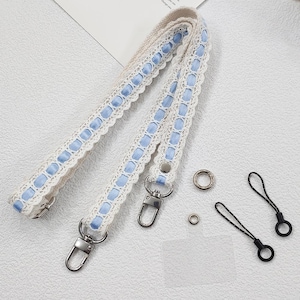 Op de afbeelding: Cr&egrave;mekleurige tasriem met blauwe lintaccenten en kanten rand. Inclusief zilverkleurige metalen sluitingen en ringen. Bevat ook twee zwarte koordlussen met zwarte ringen. Een delicaat en stijlvol accessoire.