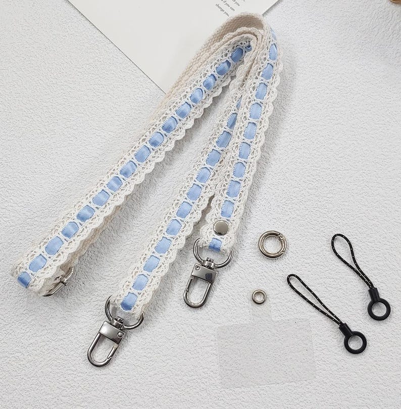 Op de afbeelding: Cr&egrave;mekleurige tasriem met blauwe lintaccenten en kanten rand. Inclusief zilverkleurige metalen sluitingen en ringen. Bevat ook twee zwarte koordlussen met zwarte ringen. Een delicaat en stijlvol accessoire.