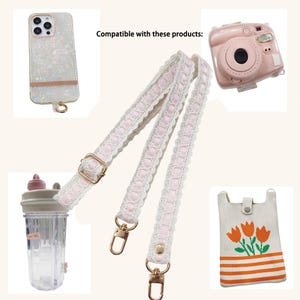 Op de afbeelding: Een witte en roze kanten band met goudkleurige hardware, afgebeeld met een roze instant camera, een telefoonhoesje, een waterfles en een kleine tas met een bloemenontwerp. De tekst "Compatible with these products:" is zichtbaar.