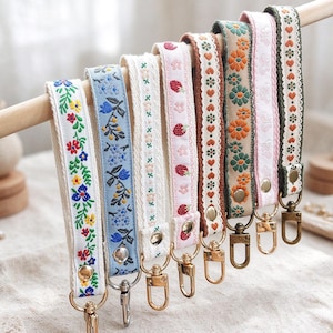 Puede incluir: Una colección de siete llaveros de tela tipo pulsera colgados de una varilla de madera. Cada correa presenta distintos patrones bordados que incluyen flores coloridas, fresas, corazones y formas geométricas, con cierres y broches de metal.