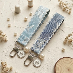 Puede incluir: Dos llaveros de tela con correas de mezclilla en azul claro y azul oscuro cubiertas con encaje floral blanco. Cada llavero incluye un mosquetón de metal plateado, un dije redondo con un pequeño lazo y una cuenta de perla blanca.