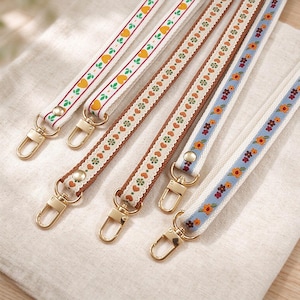 May include: Five fabric wristlet straps featuring decorative woven ribbons with floral and heart patterns. Each strap includes a gold-tone metal lobster clasp attached with a gold-tone rivet. The designs include yellow hearts, orange hearts, and colourful flowers on white, brown, and light blue backgrounds.