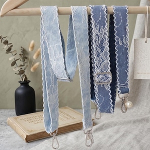 May include: Three denim bag straps with white lace trim hanging on a wooden rack. The straps vary in shades of blue and have silver-tone hardware. One strap has a pearl accent. The background includes a vase and a book.