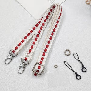 Op de afbeelding: Cr&egrave;mekleurige, gehaakte tasriem met rode lintaccenten. De riem heeft zilverkleurige metalen sluitingen en een gesp. Extra kleine zilveren ringen en zwarte koordlussen zijn ook inbegrepen.