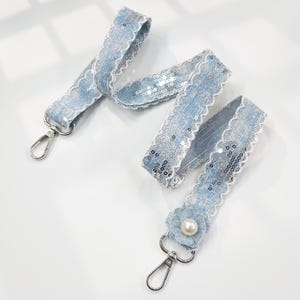 May include: Two light blue denim lanyards with silver sequin accents and white lace trim. One lanyard features a small denim flower with a pearl center. Each lanyard has a silver metal clasp.