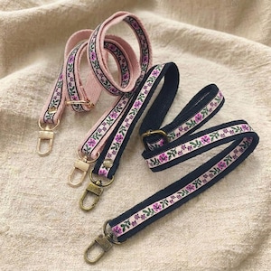 May include: Two adjustable bag straps, one in light pink and one in dark navy blue, each featuring a decorative ribbon trim with a repeating pattern of small purple flowers and green leaves, finished with gold-toned metal swivel lobster clasps.