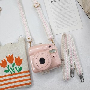 Op de afbeelding: Een roze camera met een bijpassende band met een wit kanten design. De camera heeft een grote lens en een flitser. Een kleine draagtas met oranje tulpen is ook zichtbaar.