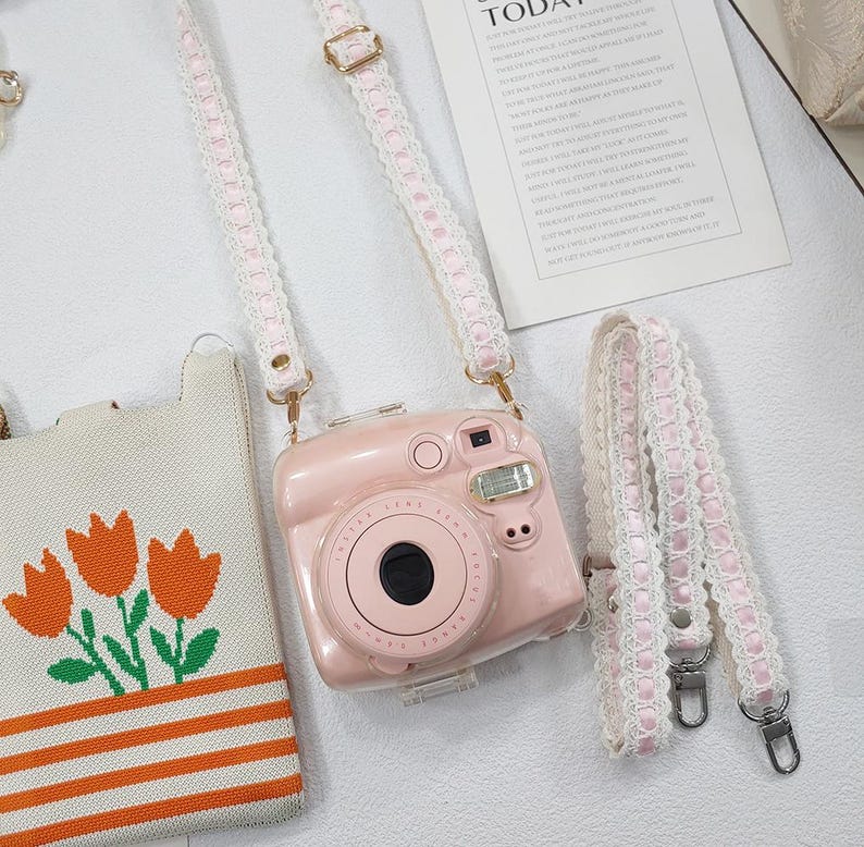 Op de afbeelding: Een roze camera met een bijpassende band met een wit kanten design. De camera heeft een grote lens en een flitser. Een kleine draagtas met oranje tulpen is ook zichtbaar.