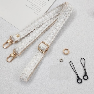 Op de afbeelding: Witte kanten tasriem met goudkleurige sluitingen en gesp. De afbeelding toont ook kleine goud- en zilverkleurige ringen en twee zwarte telefoonbedels met lussen.