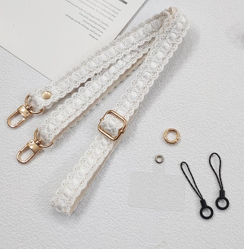 Op de afbeelding: Witte kanten tasriem met goudkleurige sluitingen en gesp. De afbeelding toont ook kleine goud- en zilverkleurige ringen en twee zwarte telefoonbedels met lussen.