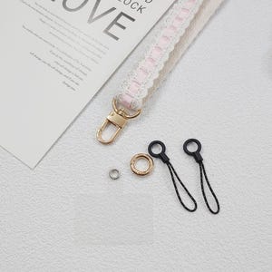 Op de afbeelding: Een close-up van een witte en roze kanten polsband met een goudkleurige sluiting en hardware. Ook afgebeeld zijn twee zwarte telefoonbedels met zwart-witte koorden, een gouden ring en een zilveren klinknagel. Het woord "LOVE" is zichtbaar.