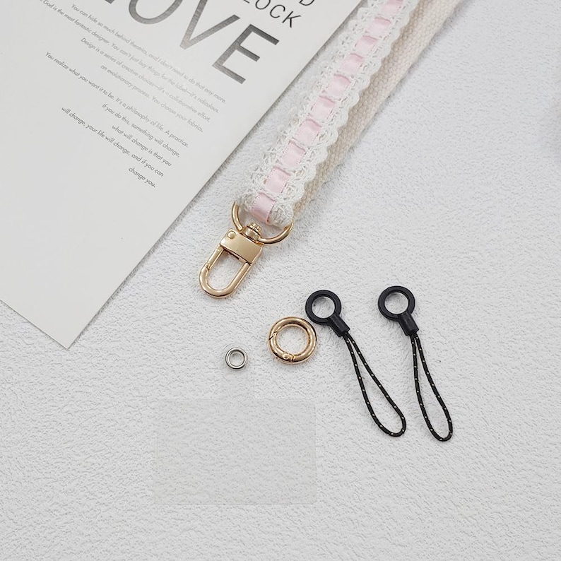 Op de afbeelding: Een close-up van een witte en roze kanten polsband met een goudkleurige sluiting en hardware. Ook afgebeeld zijn twee zwarte telefoonbedels met zwart-witte koorden, een gouden ring en een zilveren klinknagel. Het woord "LOVE" is zichtbaar.