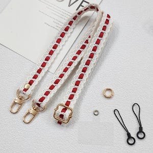Op de afbeelding: Cr&egrave;mekleurige tasriem met rode accenten en goudkleurige hardware. De riem heeft een kanten ontwerp met rode stof. Inclusief gouden ringen en zwarte koordlussen.