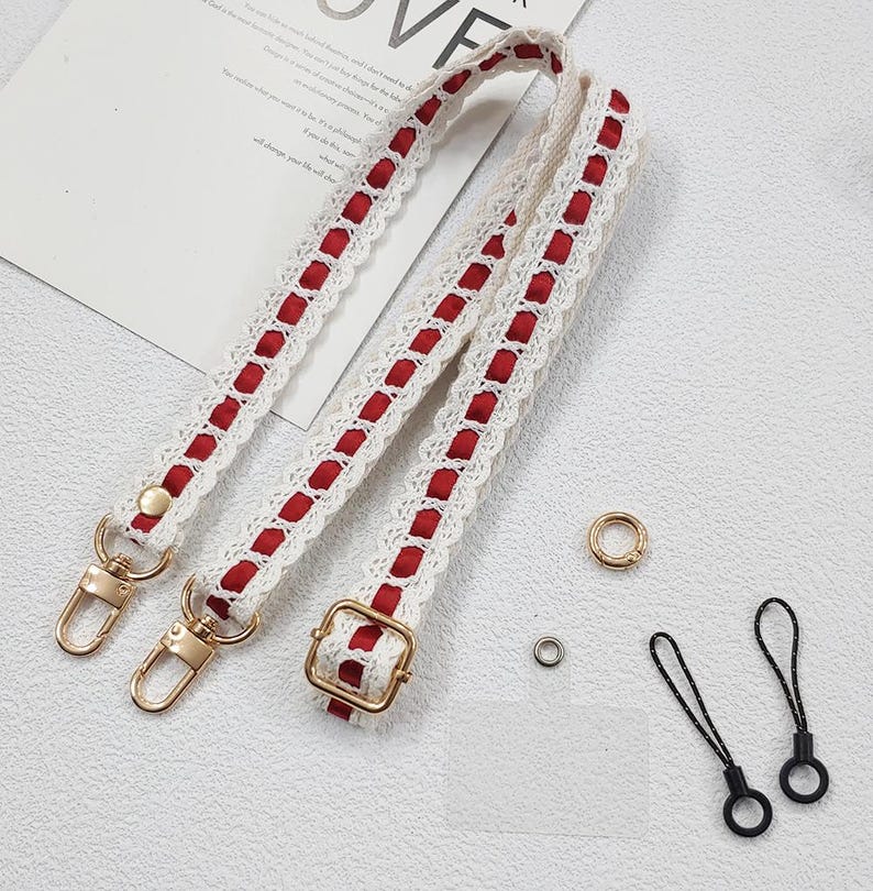 Op de afbeelding: Cr&egrave;mekleurige tasriem met rode accenten en goudkleurige hardware. De riem heeft een kanten ontwerp met rode stof. Inclusief gouden ringen en zwarte koordlussen.