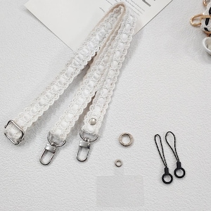 Op de afbeelding: Witte kanten band met zilverkleurige metalen bevestigingen, inclusief sluitingen en D-ringen. Kleine metalen ringen en twee zwarte lusbevestigingen zijn ook aanwezig. Het ontwerp is delicaat en vrouwelijk.
