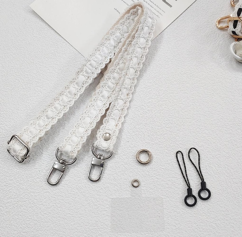 Op de afbeelding: Witte kanten band met zilverkleurige metalen bevestigingen, inclusief sluitingen en D-ringen. Kleine metalen ringen en twee zwarte lusbevestigingen zijn ook aanwezig. Het ontwerp is delicaat en vrouwelijk.
