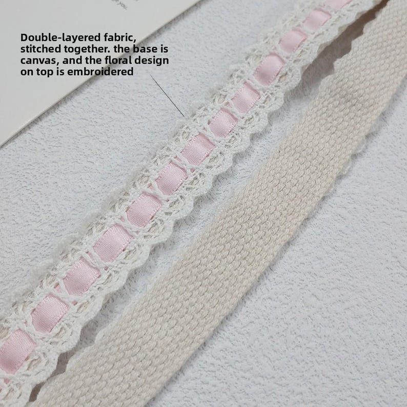 Op de afbeelding: Close-up van een dubbellaagse stof met een witte kanten rand en roze lint. De basis is canvas en het bloemmotief is geborduurd. De tekst in de afbeelding luidt: "Dubbellaagse stof, aan elkaar gestikt. De basis is canvas en het bloemmotief erboven is geborduurd."