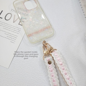 Op de afbeelding: Een doorzichtige telefoonhoes met een parelmoer design en een roze accent, samen met een wit en roze kanten telefoonriempje met goudkleurige hardware. Tekst op de afbeelding luidt "AROUND THE CLOCK" en "LOVE".