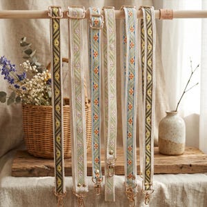 May include: A display of six decorative bag straps with various woven patterns and gold-colored hardware. The straps are cream-colored with designs in shades of green, yellow, brown, and orange. They hang from a wooden rod.