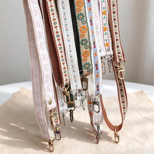 May include: A collection of hanging bag straps featuring various woven patterns, including pink butterflies, brown hearts, orange flowers, and blue floral designs. Each strap is constructed from fabric webbing and finished with gold-toned metal swivel lobster claw clasps.
