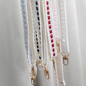 Op de afbeelding: Close-up van verschillende decoratieve riemen met goudkleurige sluitingen. De riemen zijn wit met kanten randen en hebben linten in verschillende kleuren, waaronder blauw, zwart, rood en roze. De riemen zijn ontworpen voor een tas of portemonnee.