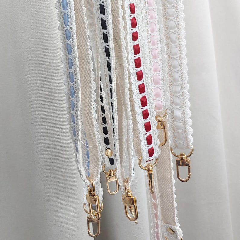 Op de afbeelding: Close-up van verschillende decoratieve riemen met goudkleurige sluitingen. De riemen zijn wit met kanten randen en hebben linten in verschillende kleuren, waaronder blauw, zwart, rood en roze. De riemen zijn ontworpen voor een tas of portemonnee.