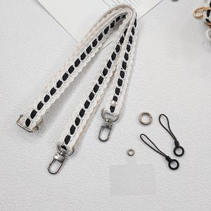 Op de afbeelding: Cr&egrave;mekleurige handtasriem met zwarte accenten en zilverkleurige hardware. De riem heeft een kanten ontwerp met zwarte stiksels. Ook inbegrepen zijn twee zwarte polsbandjes en metalen ringen.