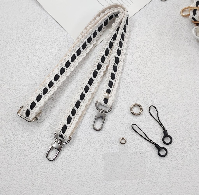 Op de afbeelding: Cr&egrave;mekleurige handtasriem met zwarte accenten en zilverkleurige hardware. De riem heeft een kanten ontwerp met zwarte stiksels. Ook inbegrepen zijn twee zwarte polsbandjes en metalen ringen.