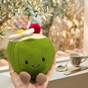 Könnte beinhalten: Ein grünes Kokosnuss-Plüschtier mit einem lächelnden Gesicht, cremefarbenem Oberteil, roter Kirsche, weißer Blume und einem blau-weiß gestreiften Strohhalm. Es hat zwei kleine, braune, abgerundete Füße. Spielzeug im tropischen Getränke-Design.