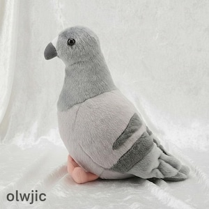 May include: A soft grey pigeon toy with a black beak and eye details. The stuffed animal has a light grey body with darker grey wing markings and pink feet. The toy is sitting on a white surface.